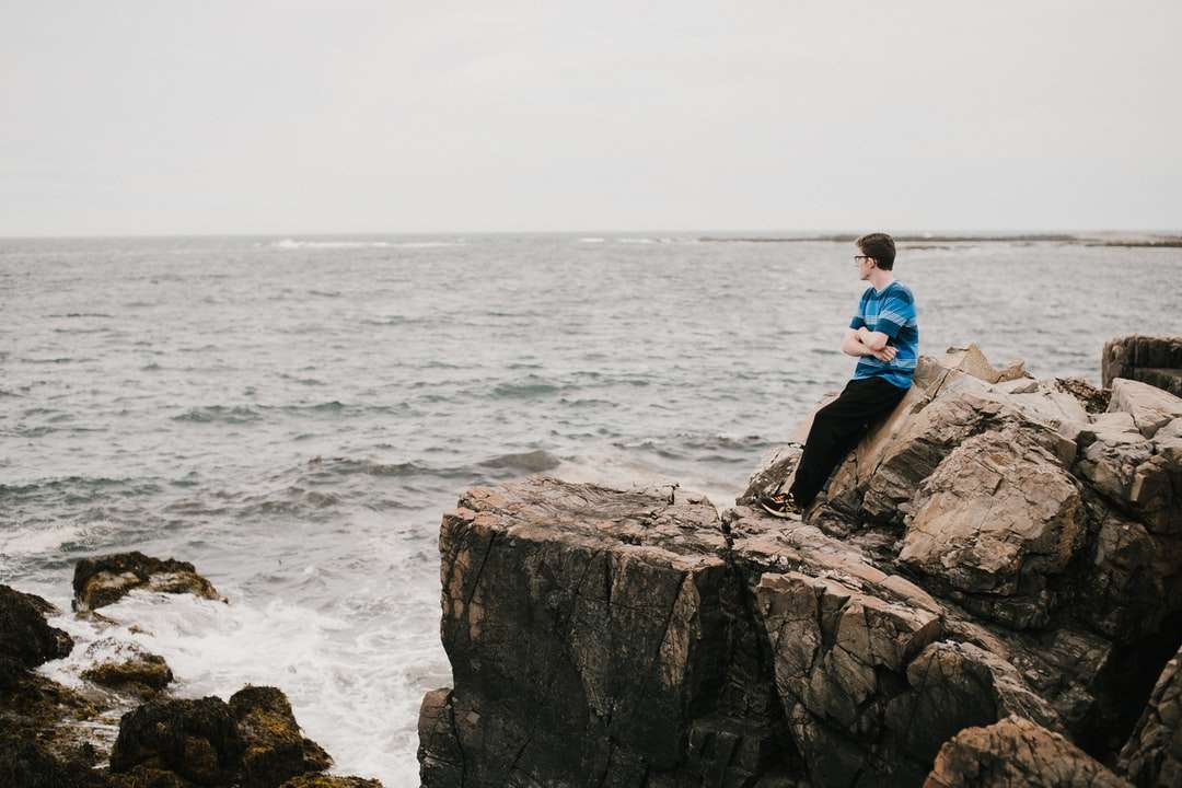 mężczyzna w niebieskiej koszuli siedzi na formacji skalnej w pobliżu morza puzzle online