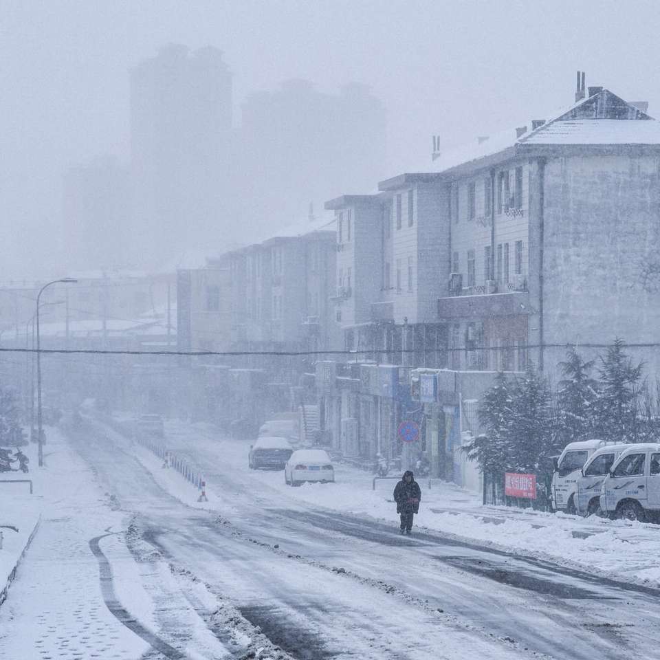 osoba idąca po zaśnieżonej drodze w pobliżu samochodów i budynków puzzle online