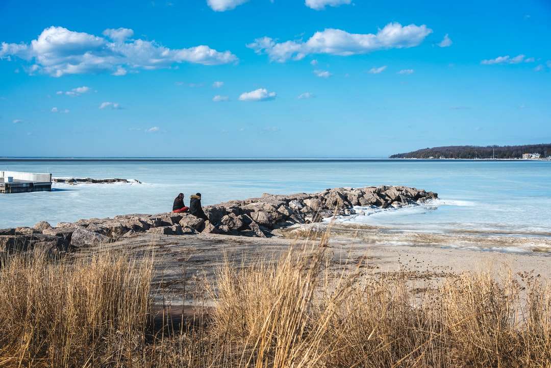 Mężczyzna siedzi na skale w pobliżu morza pod błękitnym niebem w ciągu dnia puzzle online