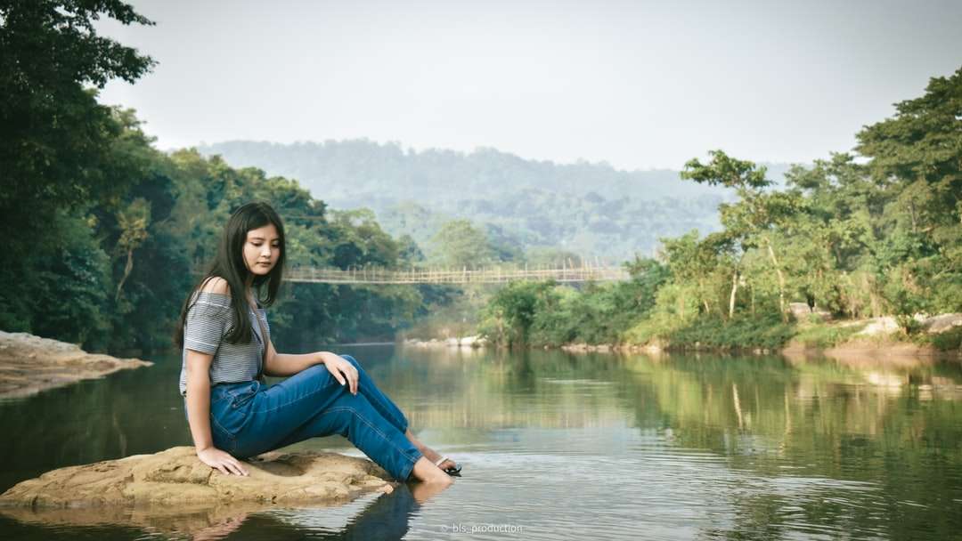 Kobieta w niebieskich dżinsach siedzi na brązowej skale w pobliżu jeziora puzzle online