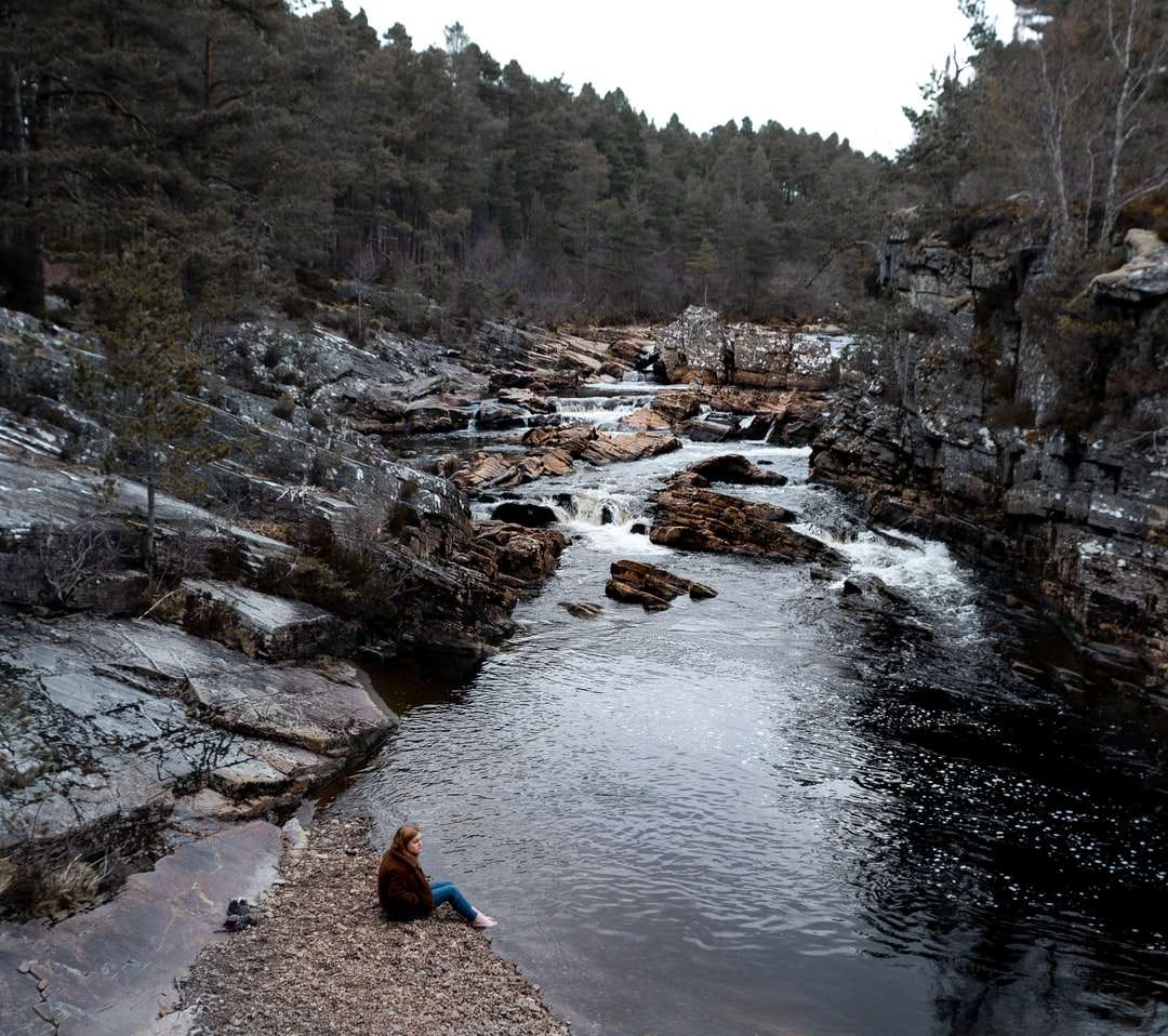 Osoba w niebieskiej kurtce siedzi na skale w pobliżu rzeki puzzle online