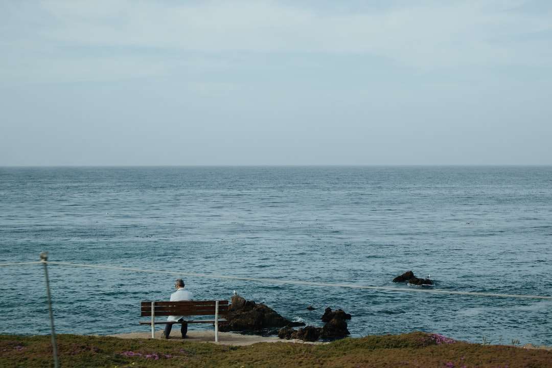 Osoba siedzi na białej drewnianej ławce w pobliżu morza w ciągu dnia puzzle online