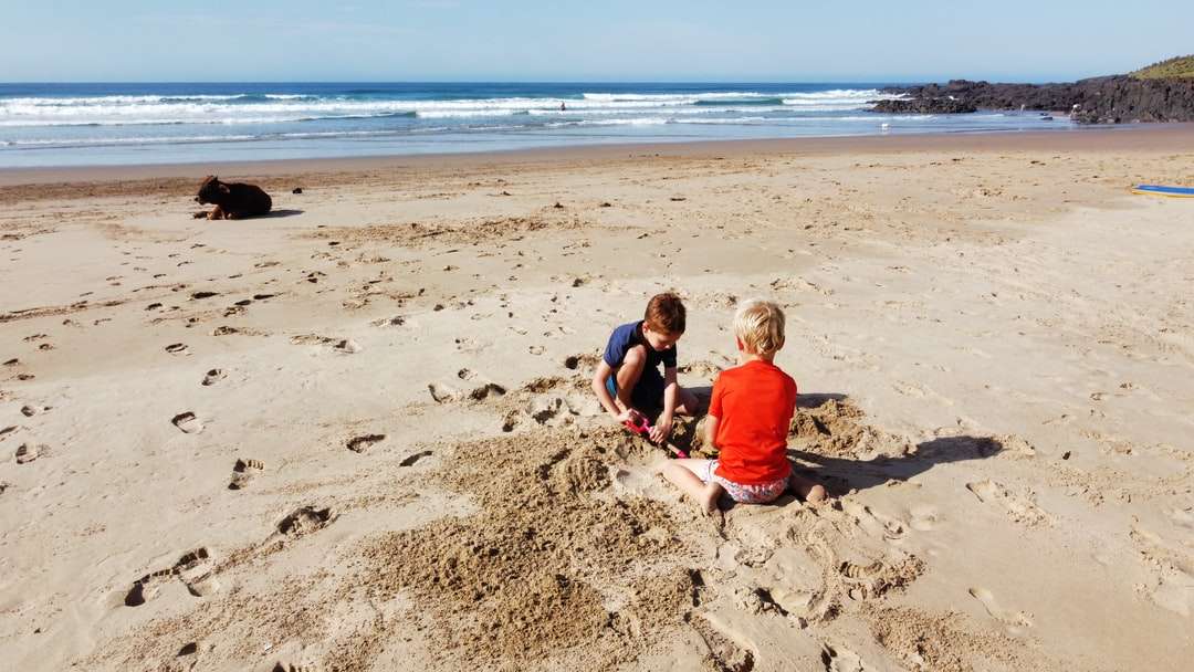 2 chłopców siedzi na brązowym piasku w pobliżu morza w ciągu dnia puzzle online
